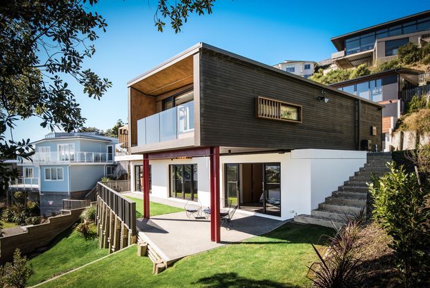 Exterior view of a contemporary multi-level hillside residence with a cantilevered dark timber-clad upper volume, white lower walls, red structural posts, extensive glazing, and a sheltered outdoor terrace. The house sits on a steep landscaped site with stairs, retaining structures, and neighbouring homes visible around it.
