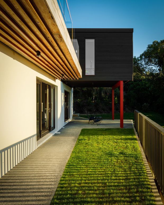 Exterior architectural photograph of a contemporary two-level house with a projecting dark horizontal-clad upper volume supported by slender red posts. The lower level has pale rendered walls, glazed doors, and a deep timber-lined soffit with exposed beams. A paved walkway, narrow lawn, and timber balustrade occupy the foreground, with mature vegetation beyond.