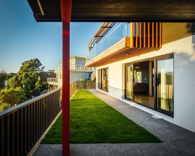 Exterior view of a contemporary multi-level residential building, showing a landscaped rooftop or elevated terrace with lawn, dark gravel paving, black metal balustrades, extensive glazing, timber-lined soffits, and a prominent red structural column. The building has projecting upper-level balconies and a clean modern facade, with surrounding greenery and neighboring structures visible beyond.