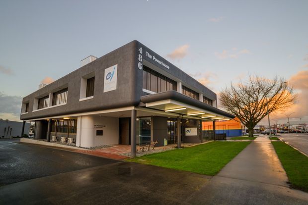 Exterior view of a contemporary two-storey commercial building at dusk, with dark textured cladding, extensive glazing, a projecting entrance canopy, and prominent business signage. The building fronts a paved sidewalk with a small lawn and street trees, and appears to be part of an urban commercial setting.