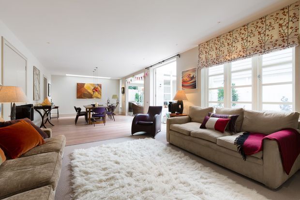 Bright, spacious open-plan living and dining area with large windows, patterned Roman blinds, neutral upholstered sofas, a leather armchair, dining table, artwork, and a soft white shag rug. The room has a refined traditional-contemporary feel with warm timber flooring and abundant natural light.