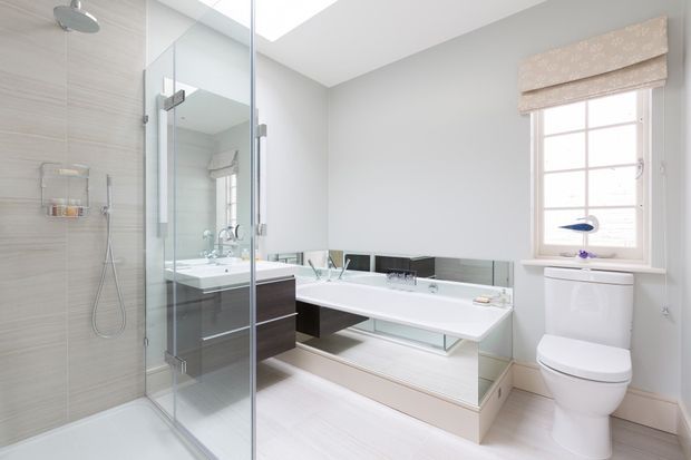 Bright, contemporary bathroom with a frameless glass walk-in shower, wall-mounted handheld shower, floating dark-wood vanity, long white integrated basin, mirrored splashback, freestanding-style toilet, pale timber-look flooring, and a window with a patterned Roman blind. A skylight adds natural illumination.