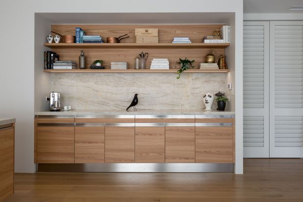 Built-in timber joinery wall with open display shelving, a pale stone feature backsplash, integrated cabinetry, and a coffee station. The warm wood cabinetry and shelving contrast with brushed metal trim and a grey painted wall, while decorative objects and books add a refined, lived-in character. White louvered doors are visible to the right.
