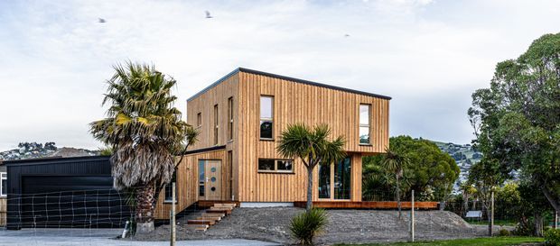 Exterior view of a modern two-storey residence with a simple rectangular form, vertical timber cladding, dark-framed windows, and a dark flat or very low-pitched roof. The house is set above a landscaped gravel frontage with timber steps, a covered garage or carport structure to the left, and palm-like planting around the entry. A wooded, hilly suburban setting is visible behind the property.