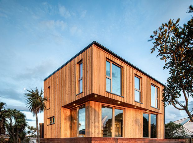 A contemporary two-storey residential house viewed from the front corner, clad in warm vertical timber boards. Large full-height windows and glazed doors punctuate the facade, with a projecting upper-floor volume forming a sheltered entry or terrace below. The building has a simple pitched roof, dark metal roof edging, and a timber deck or low platform at ground level. Coastal-style planting and neighbouring homes are visible around the exterior.