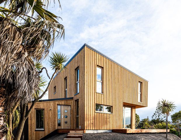 Exterior view of a contemporary two-storey house clad in vertical natural timber boards. The angular gabled form includes tall narrow windows, a projecting upper level, a timber entry door, and a small deck or landing. Coastal-looking vegetation surrounds the home, with bright daylight and glimpses of open landscape beyond.