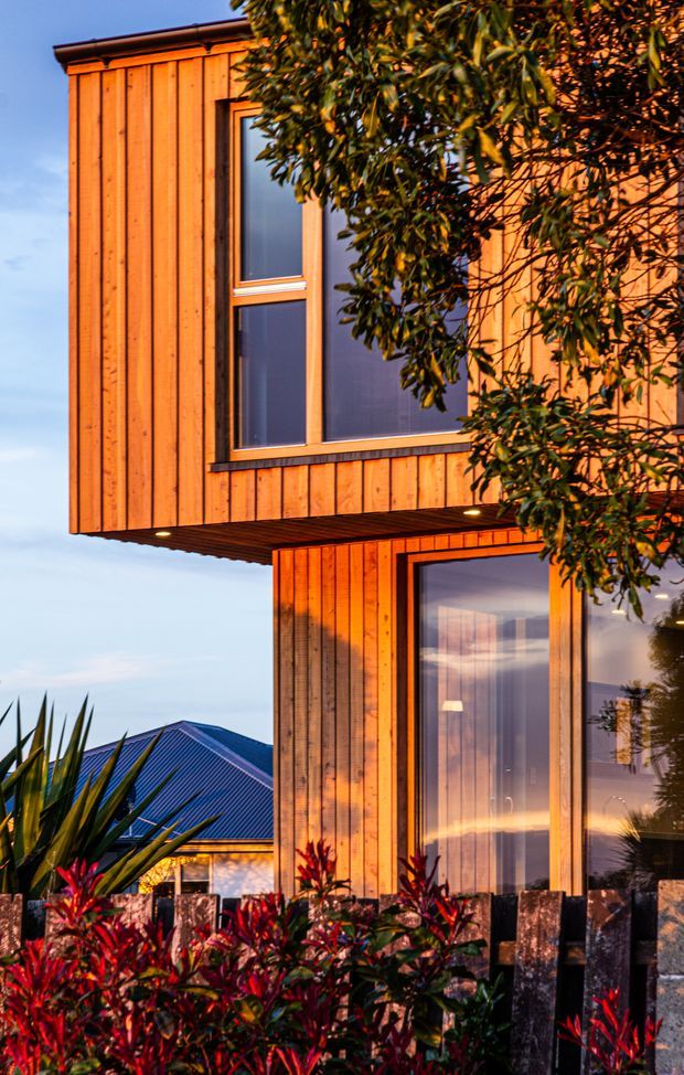 Exterior architectural photograph of a contemporary two-storey home with projecting upper-floor volumes, vertical warm timber cladding, dark window frames, and large glazed openings. The building is partially screened by mature foliage and colourful landscaping, with a neighbouring pitched metal roof visible in the background. Warm low-angle sunlight gives the facade an orange glow.