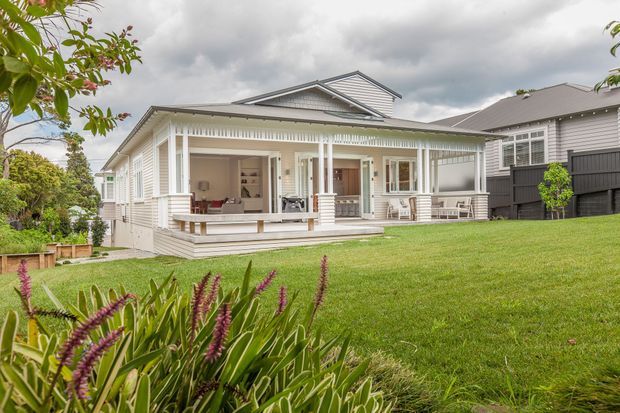 Exterior view of a light-painted, single-storey weatherboard house with a broad covered veranda, decorative balustrade and posts, gabled roof, and multiple timber-framed windows and doors. The house sits in a landscaped suburban garden with a large lawn and planting, under an overcast sky. A darker neighbouring weatherboard house is visible to the right.