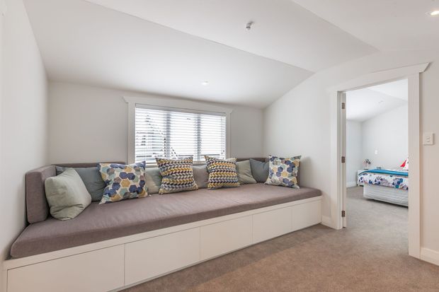 Bright, compact sitting room with a long built-in window seat and integrated white storage cabinetry beneath it. The seat is upholstered in muted mauve-grey fabric and layered with patterned and plain cushions. A wide window with horizontal blinds provides natural light, while an open doorway reveals part of an adjacent bedroom. Neutral walls and beige carpet create a clean, comfortable interior.
