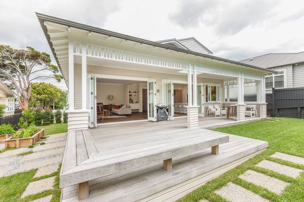 Bright exterior view of a single-storey coastal-style home with a broad covered veranda, white painted timber detailing, open bi-fold doors, and a large weathered timber deck with stepped seating. The house has a pitched metal roof with a glazed roof section, horizontal weatherboard cladding, decorative balustrade screening, and a neatly landscaped lawn and garden. A living room and dining area are visible through the open doors.
