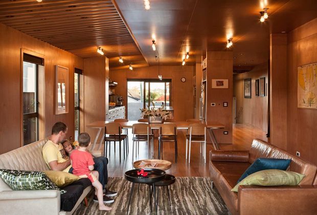 A warm, open-plan mid-century-inspired interior combining a living room, dining area, and compact kitchen. Timber wall panelling and ceiling details create a rich amber backdrop, with a central dining table and bar stools, leather sofas, a patterned rug, and track-style ceiling lighting. Large glazed doors and windows provide views toward the outdoors.
