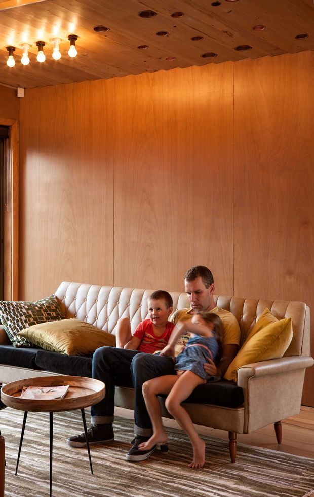 Warmly lit mid-century-inspired living room with extensive honey-toned timber wall and ceiling panelling, a tufted cream sofa, patterned and mustard cushions, a round timber coffee table, and a striped area rug. A parent and two children are seated on the sofa. Recessed ceiling lights and exposed bulb fixtures create an intimate, inviting atmosphere.