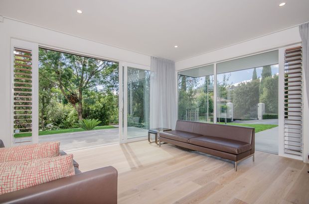 Bright contemporary living room with pale timber flooring, white walls and ceiling, a brown leather sofa, and patterned upholstered armchairs. Floor-to-ceiling sliding glass doors and windows open the room visually to a landscaped garden, with white plantation shutters and sheer curtains framing the glazing.