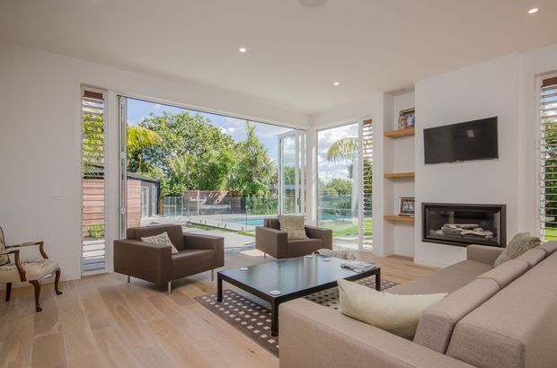 Bright contemporary living room with light timber flooring, neutral upholstered seating, a dark coffee table, built-in shelving, wall-mounted television and fireplace. Floor-to-ceiling glazed corner doors and windows open the room visually to a landscaped outdoor area and swimming pool, with louvred window shutters providing filtered light.