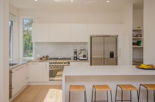 Bright contemporary kitchen with a large white island and integrated breakfast bar, pale timber flooring, white handleless cabinetry, stone-look splashback, stainless-steel appliances, and open shelving. A large window brings in natural light and views of greenery.