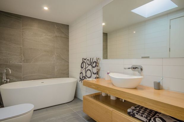 Bright contemporary bathroom featuring a freestanding white bathtub, wall-mounted toilet, floating timber vanity with a white vessel basin, large frameless mirror, white horizontal wall tiles, grey stone-look floor and feature wall tiles, and a skylight.