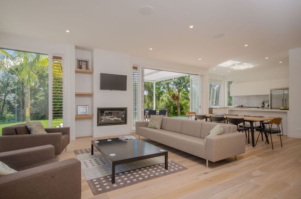 Bright open-plan contemporary living, dining and kitchen interior with pale timber flooring, white walls, recessed lighting and extensive glazing opening to greenery. The living area features a large neutral sectional sofa, coffee table and armchairs, with built-in shelving, wall-mounted television and fireplace. A dining table and island kitchen are visible in the background.