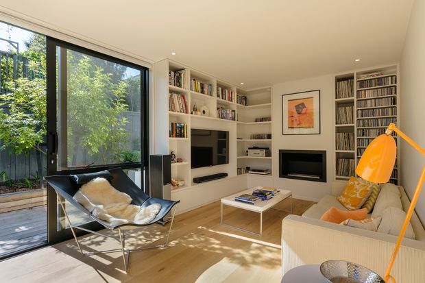 A bright, compact contemporary living room with extensive built-in white shelving and cabinetry, filled with books and records. The room includes a wall-mounted television, a separate fireplace or media feature, a cream sofa, black sling chair, coffee table, and a vivid orange floor lamp. Large black-framed sliding glass doors open to a timber deck and leafy outdoor area, bringing in abundant natural light.