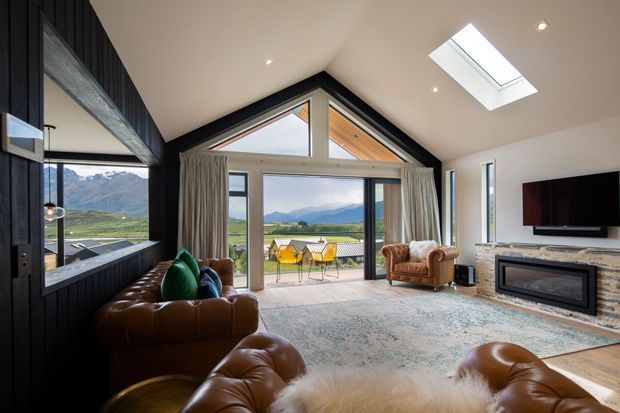 A spacious contemporary living room with a dramatic vaulted gable ceiling, exposed timber detailing, skylight, and expansive glazed doors opening to a mountain and rural landscape. The room features dark vertical wall panelling, a large patterned rug, leather sofas and armchairs, a fireplace, and a wall-mounted television.