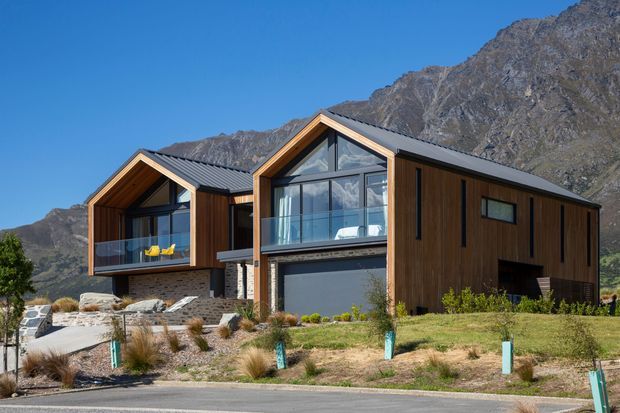 Exterior view of a contemporary mountain residence composed of two connected gabled volumes. The home combines dark vertical timber cladding, charcoal standing-seam metal roofing, extensive glazed gable ends with balconies, and a dark masonry base. It sits on a landscaped sloping site with the rugged mountain backdrop clearly visible.