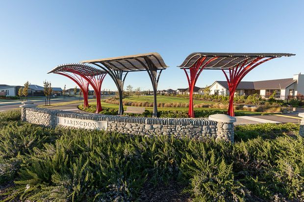 A landscaped residential development entrance featuring a curved, sculptural steel canopy structure supported by red and dark branching columns. A low rounded-stone retaining wall bears the “Faringdon” name, with paved walkways, garden planting, and low-rise homes visible in the background under a clear sky.