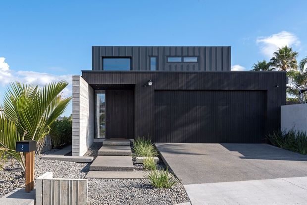 Front exterior view of a contemporary two-storey residence with a dark vertically ribbed facade, integrated double garage, recessed entry, narrow modern windows, and a minimalist gravel-and-planting landscape.