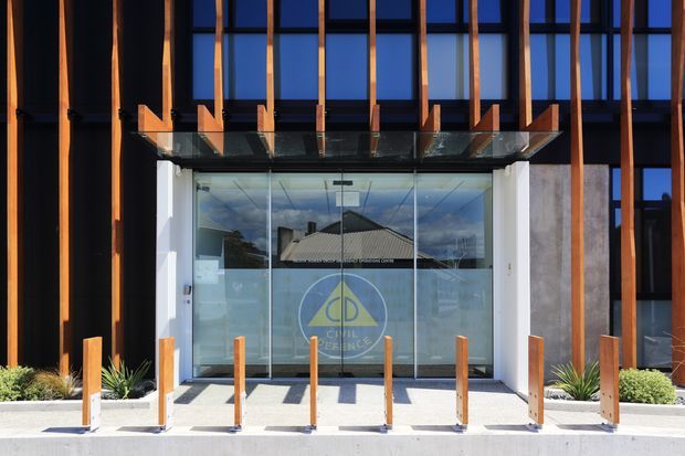 Contemporary commercial building entrance with a glazed vestibule framed by white structural walls, a projecting glass canopy, and a dark facade articulated with vertical timber-toned fins. Tall blue-tinted glazing extends above the entry. Timber bollards line the concrete approach, with low planting beds at the sides. A circular logo is visible on the entrance glazing.