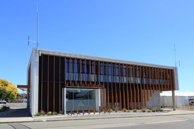 A contemporary low-rise commercial building viewed from the street, with a crisp white rectangular envelope, extensive glazing, and a prominent facade screen of closely spaced vertical timber fins. The upper level projects slightly over the glazed entrance, creating a sheltered entry and strong horizontal composition. Landscaping is minimal, with a paved verge, small planted beds, and clear blue sky.