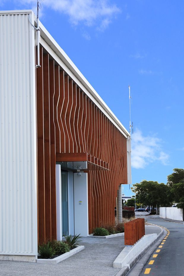 Exterior view of a contemporary commercial or institutional building with a steeply sloping gable-like facade. The facade combines white corrugated metal cladding with a warm timber vertical-slat screen that extends over the entrance, creating a strong geometric rhythm. A paved pedestrian edge, integrated planters, bollards, and a marked street are visible under a bright blue sky.