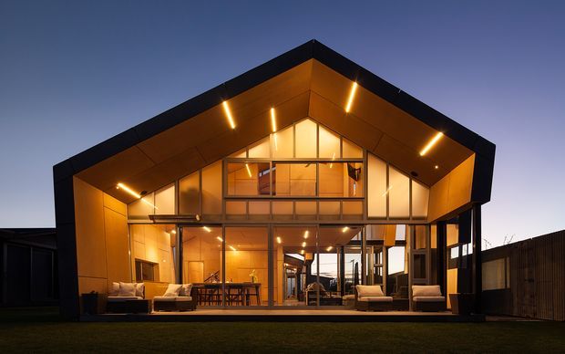 Dusk exterior view of a contemporary single-storey gable-roof home with a dramatically illuminated timber-lined porch and large floor-to-ceiling glazed facade. The open front reveals a warm living and dining area, with an upper-level mezzanine visible beneath the pitched roof. Dark cladding frames the roof and facade, while outdoor seating and a lawn occupy the foreground.