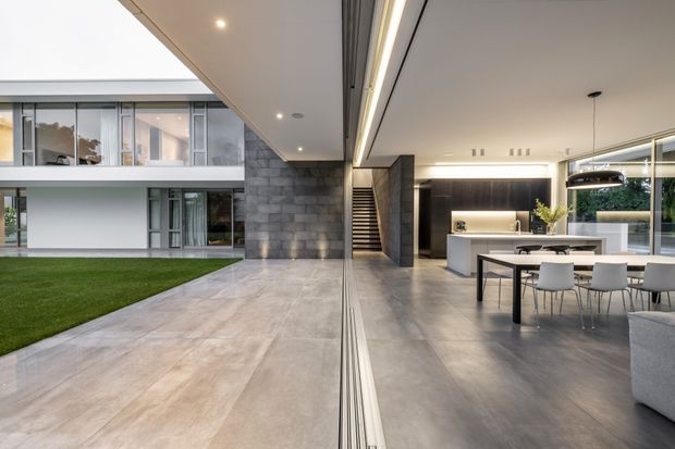A spacious contemporary open-plan living, dining and kitchen area opening onto a manicured courtyard lawn. The room features polished concrete-look flooring, dark stone-clad walls, extensive floor-to-ceiling glazing, a minimalist kitchen with island, a dining table, and a stairway leading to an upper level. Recessed linear lighting and a large pendant add a refined modern finish.