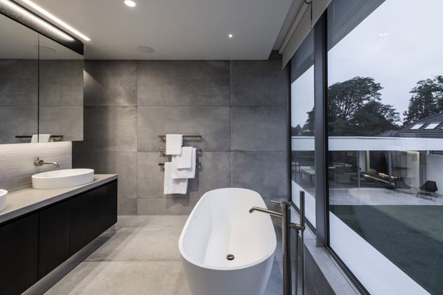 Contemporary bathroom with a freestanding white bathtub beside full-height glazing, a floating dark vanity with vessel basin, large-format grey wall and floor tiles, integrated lighting, and views to an outdoor courtyard.