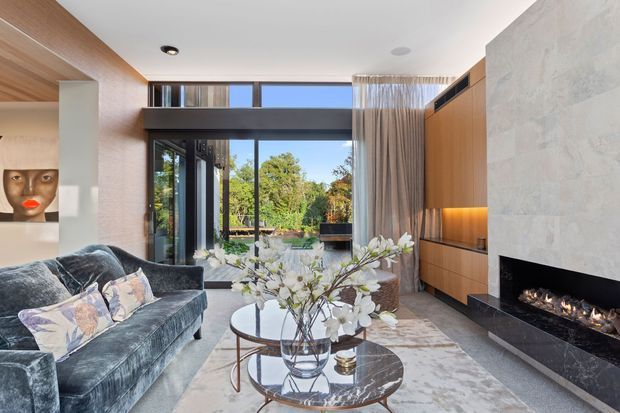 Contemporary living room with a velvet sectional sofa, round nesting coffee tables, floor-to-ceiling glazing, timber built-in cabinetry, and a large pale stone fireplace surround with a linear gas fire. The room opens to a landscaped outdoor area and features layered sheer curtains, recessed lighting, and a neutral grey, beige, and timber palette.
