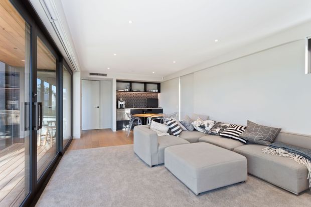 Bright, open-plan contemporary living room with a large light-grey sectional sofa and ottoman, patterned cushions, and a pale area rug. Full-height dark-framed sliding glass doors line one side, opening toward an outdoor area, while a compact kitchen with dark cabinetry and a tiled splashback is visible at the rear. The room has a clean white ceiling with recessed downlights and warm timber flooring near the kitchen.