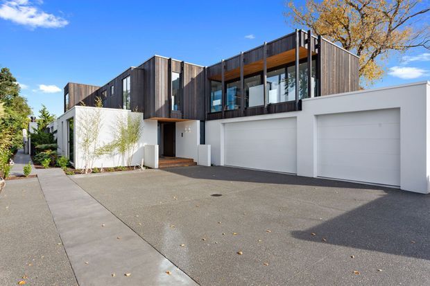 Modern multi-level residential house viewed from the street, featuring dark vertical timber cladding, crisp white rendered walls, extensive upper-level glazing, projecting balconies, and an integrated double garage. A paved driveway and pedestrian path lead to the recessed entry, with mature landscaping around the site.