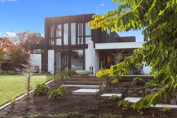Exterior photograph of a contemporary multi-storey residence set within a landscaped garden. The house features a dark vertical timber-clad upper volume, extensive floor-to-ceiling glazing, white rendered lower walls, and a projecting flat-roofed entrance or outdoor living canopy. A stepped stone garden path and retaining wall lead toward the building.