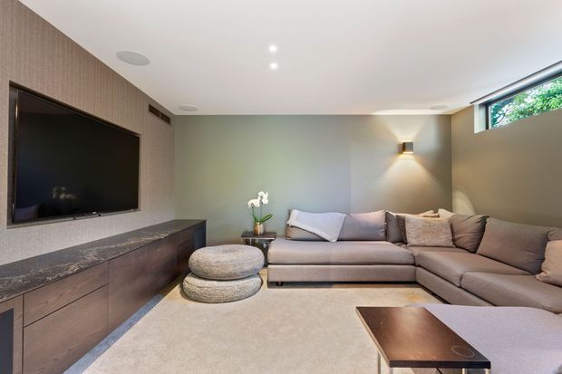 A refined contemporary living room featuring a large L-shaped upholstered sectional, layered round ottomans, a wall-mounted television, and a low timber entertainment cabinet with a dark stone benchtop. The room has muted olive-grey walls, soft beige carpet, recessed ceiling lights, and a warm wall sconce, with a narrow high-level window bringing in greenery and natural light.