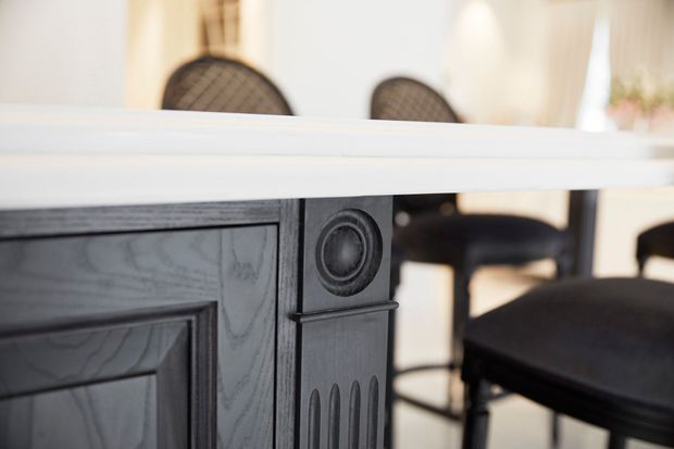 Close-up interior view of a dark painted timber kitchen island or built-in joinery featuring traditional panelled detailing and a decorative circular carved motif. A pale benchtop or counter surface spans the foreground, with dark upholstered dining or bar chairs visible in the softly lit background.