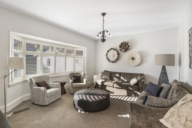 Bright, comfortably furnished living room with a large bay window fitted with white plantation shutters, neutral carpeting and walls, multiple upholstered armchairs and sofas, a round tufted ottoman, decorative wall art, floor lamps, and a central hanging chandelier.