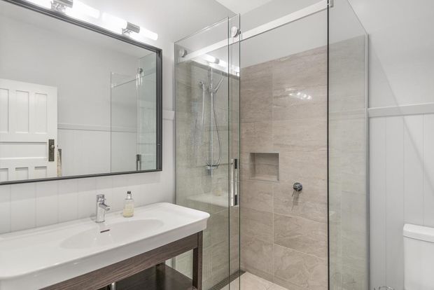 Bright, compact bathroom featuring a large vanity with white basin, wall-mounted mirror, frameless glass shower enclosure, beige stone-look tiled shower walls, white vertical wall panelling, and a toilet. The space has a clean, traditional-contemporary fit-out with chrome fixtures and timber-toned vanity cabinetry.