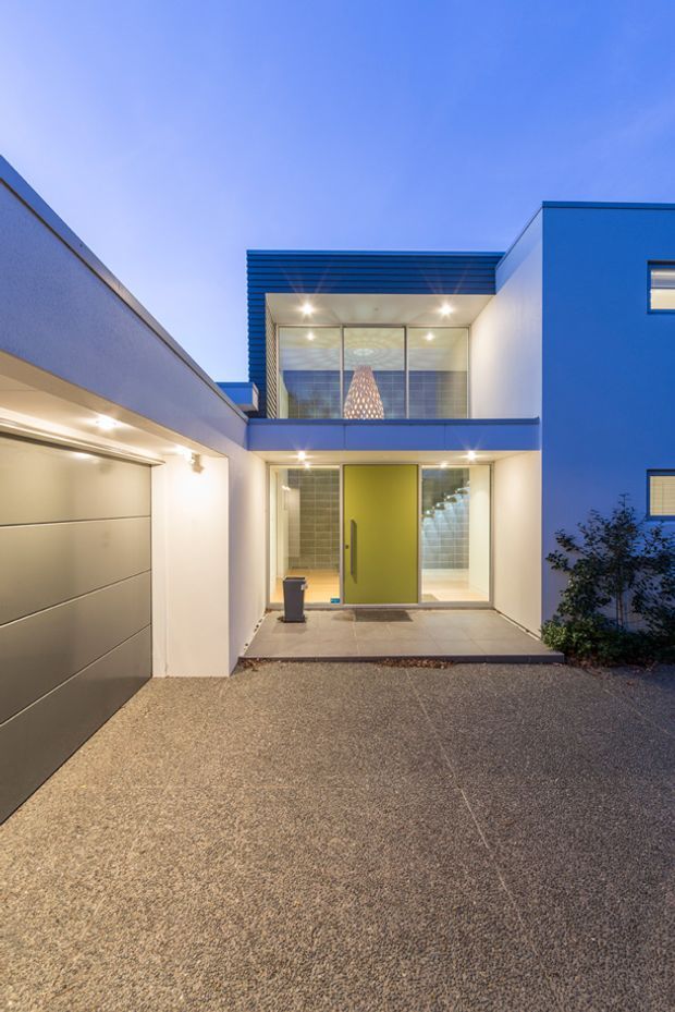 Contemporary two-storey residential facade viewed from the driveway at dusk. The home features clean white rendered walls, bold blue exterior cladding and trim, extensive glazed entry areas, a large garage door, a sheltered front entrance with a bright lime-green door, and modern exterior lighting. A small landscaped planting area borders the right side.