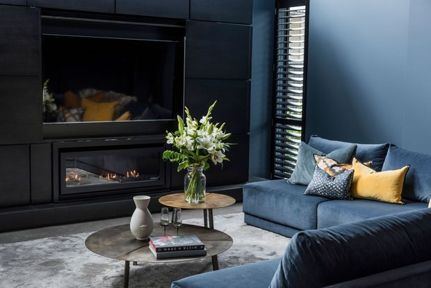 A sophisticated dark-toned living room with a built-in black feature wall incorporating a television and linear fireplace. A deep blue sectional sofa with patterned and mustard cushions faces layered round coffee tables, while a large grey rug, floral arrangement, and louvred window add texture and contrast.