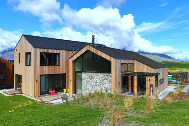Exterior view of a contemporary multi-storey residence with timber-clad walls, dark standing-seam pitched roofs, large glazed openings, and a prominent central gabled entry volume faced in rough natural stone. The house sits in a landscaped rural or semi-rural setting with mountains visible beyond.