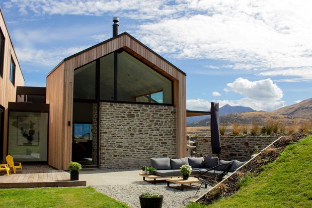 Exterior view of a contemporary gabled house set in a mountainous landscape. The facade combines vertical timber cladding, extensive dark-framed glazing, and a prominent rough stone masonry wall. A timber deck and gravel outdoor seating area with lounge furniture occupy the foreground, with a sloping grass bank and distant hills beyond.