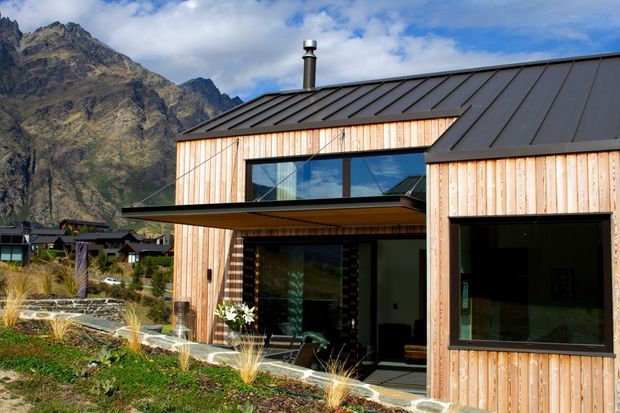 Contemporary single-storey mountain home with a dark standing-seam metal roof, vertical light timber cladding, large black-framed glazing, and a projecting covered terrace. The house is set against a dramatic alpine mountain backdrop with landscaped planting and stone edging.