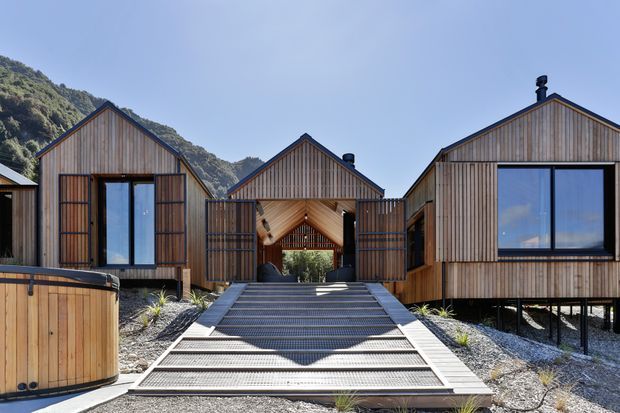 Exterior view of a contemporary cluster of timber-clad gabled buildings set on a sloping, mountainous site. The central covered entry is approached by broad stepped decking and a walkway, with dark-framed glazing, vertical timber screens, raised structures, and a small timber outdoor spa or tub in the foreground.