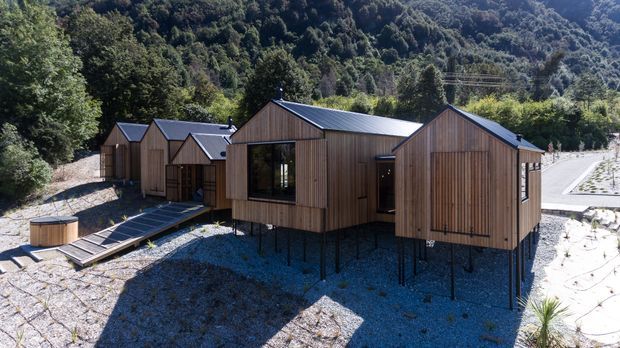 Exterior view of a group of compact timber-clad buildings on slender raised supports, arranged on a wooded, sloping site. The structures have repeated pitched gable roofs, dark metal roofing, large glazed openings, and connecting timber walkways. A forested hillside and gravelled landscape surround the development.