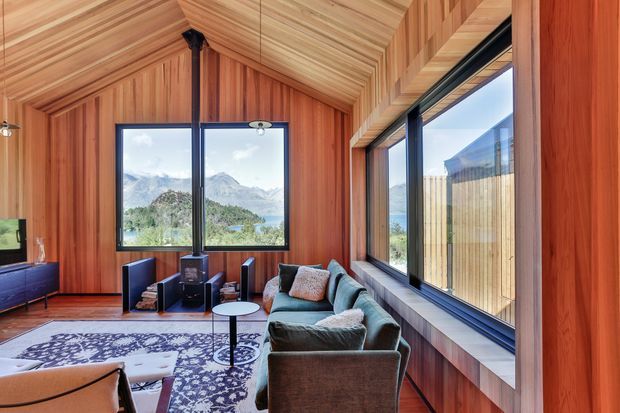 A warm, timber-lined living room with a sloping wood-clad ceiling and extensive black-framed windows overlooking a mountainous landscape. The room contains a dark sectional sofa, patterned rug, small round coffee table, compact wood-burning stove, and low media cabinetry. The strong natural materials and panoramic glazing create a cozy yet expansive retreat-like atmosphere.
