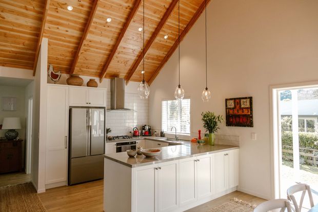 Bright open-plan kitchen with white Shaker-style cabinetry, stainless-steel refrigerator and appliances, a central island with an integrated sink, and pendant lighting. The space features a dramatic pitched ceiling clad in warm timber boards with exposed dark timber beams, timber flooring, and large windows and glazed doors providing abundant natural light.
