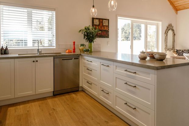Bright, warm kitchen with white shaker-style cabinetry arranged around an L-shaped layout and a large island or peninsula with a stone benchtop. Timber flooring, white walls, pendant lights, extensive windows and glazed doors create an airy connection to the outdoors. Decorative accessories and a mirror add a traditional, homely character.